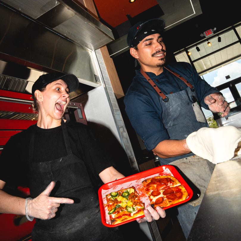 Two chefs in a kitchen, one holding a tray of pizza slices, the other stretching pizza dough.