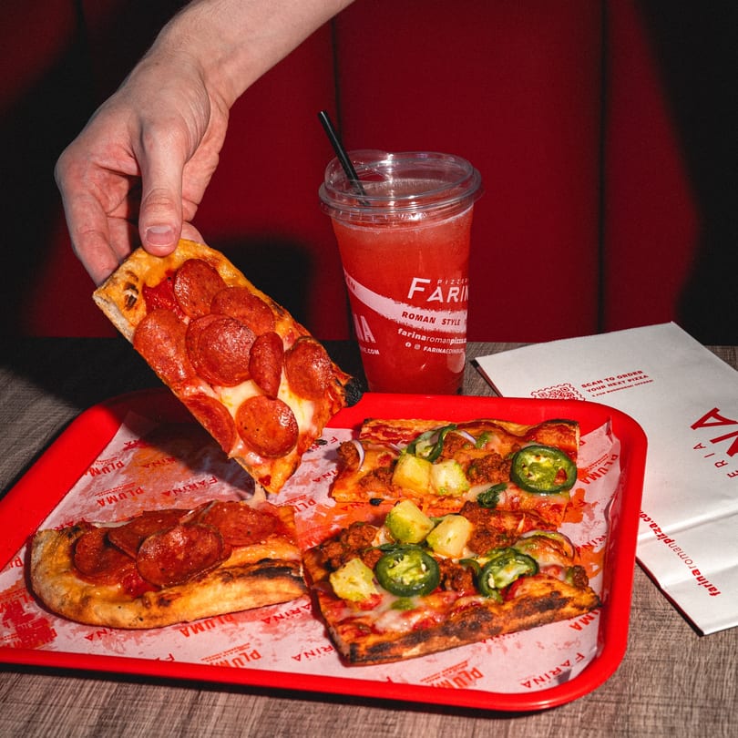 A hand lifts a slice of pepperoni pizza from a red tray with other pizza slices and a drink.